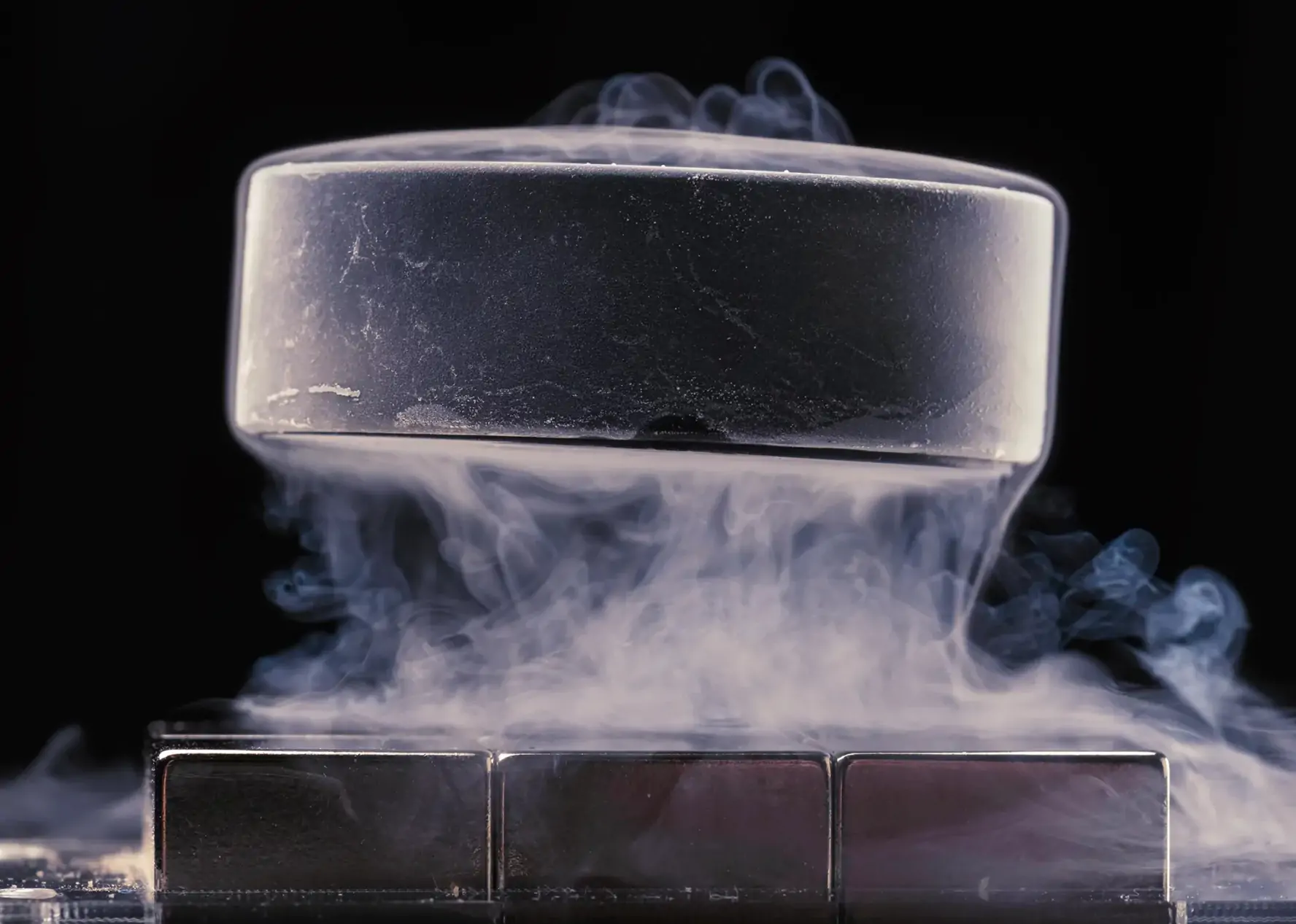 A squat dark cylinder of superconductive material levitating in air, surrounded by a wispy fog like substance from liquid nitrogen cooling the air